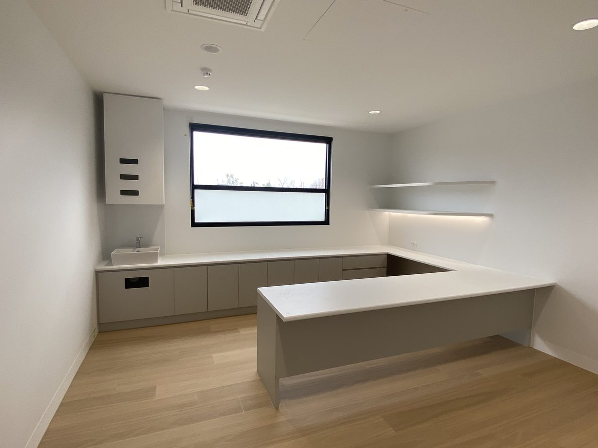 Empty consulting room with a wrap-around benchtop over drawer and cupboard joinery, a basin under a tall cabinet and two floating shelves, Bayside.