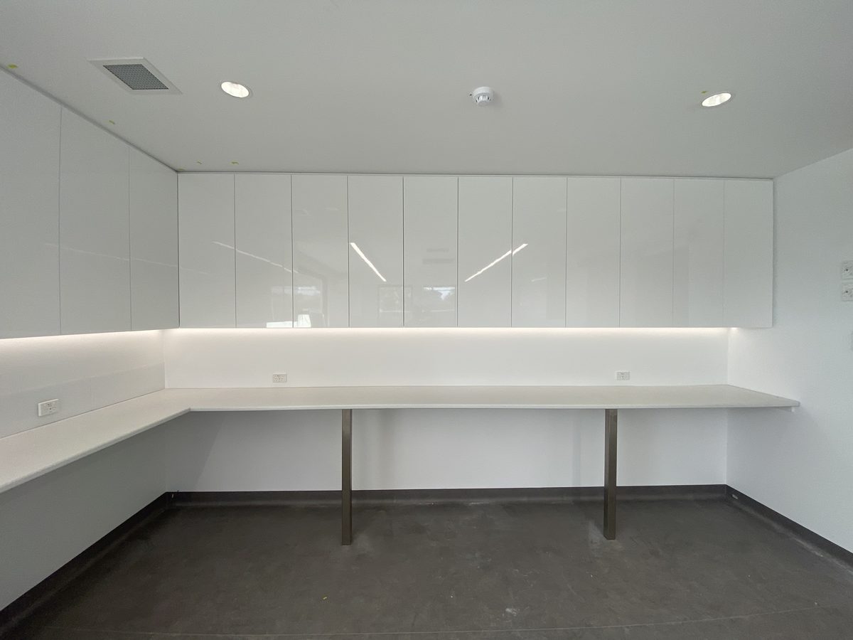 Joinery bench run with gloss overhead cupboards, under-cupboard lighting and a benchtop on steel legs in a medical centre, Bayside.