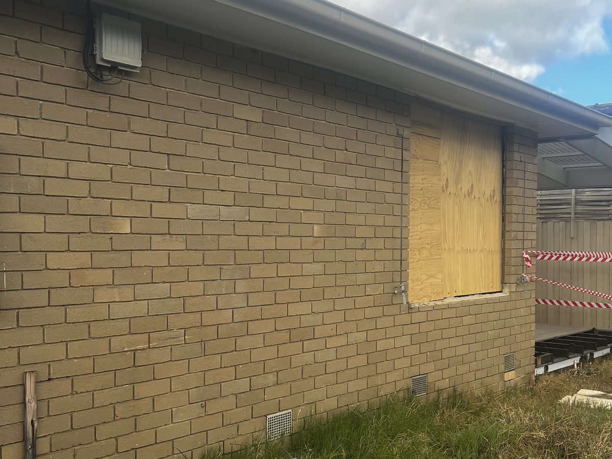Side elevation of the original cream brick house with a window opening sheeted in plywood and barrier tape across the corner as the works begin, Melbourne’s south-east.