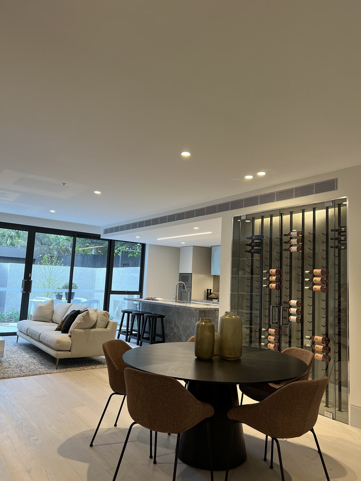 Finished open-plan living, dining and kitchen area with a stone island bench and a glazed wine display wall, Bayside.